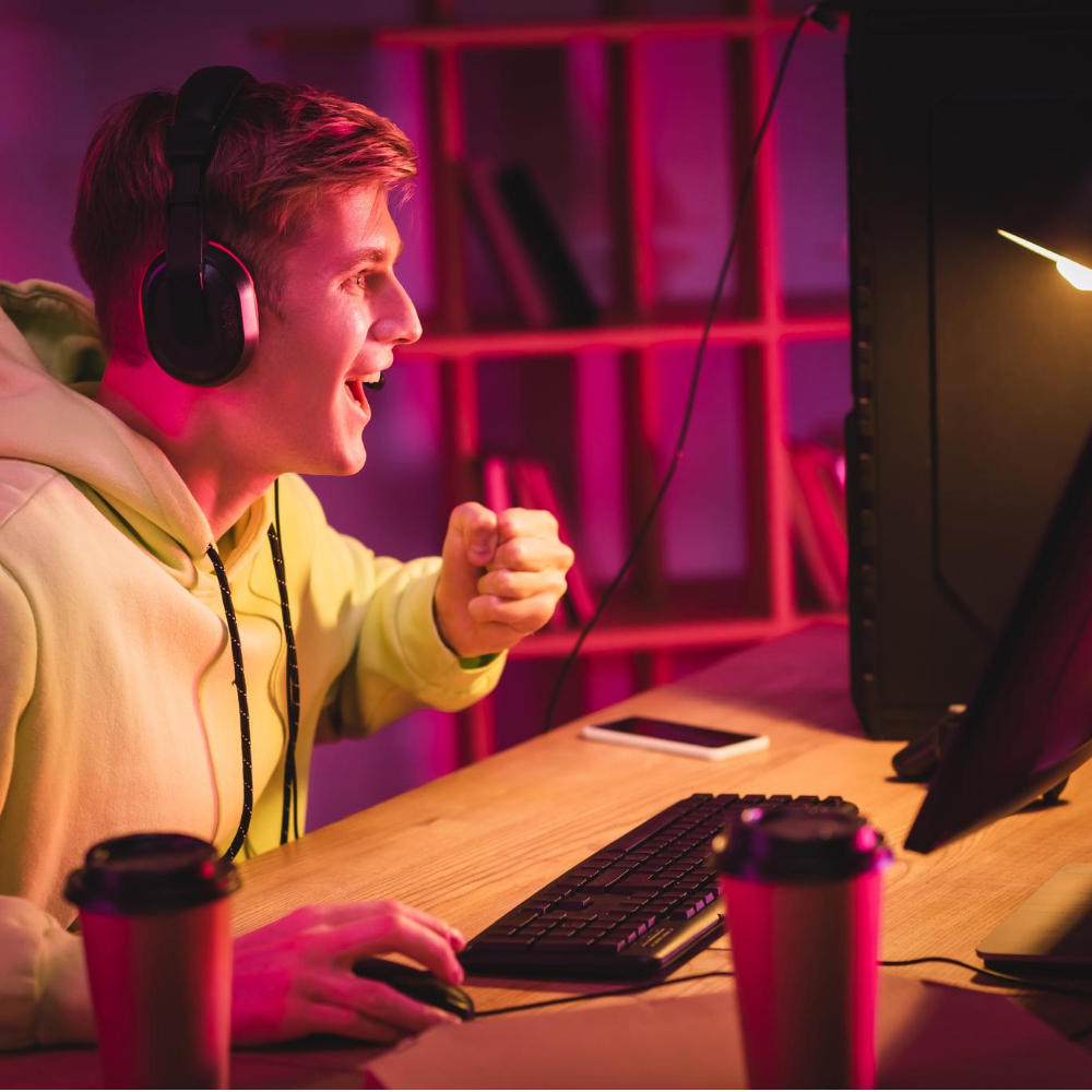 A young person wearing headphones and a hoodie sits at a desk, looking excitedly at a computer screen, surrounded by coffee cups, a keyboard, and a phone—ready to dive into gaming customer service or video game customer support tasks. A young person wearing headphones and a hoodie sits at a desk, looking excitedly at a computer screen, surrounded by coffee cups, a keyboard, and a phone—ready to dive into gaming customer service or video game customer support tasks.