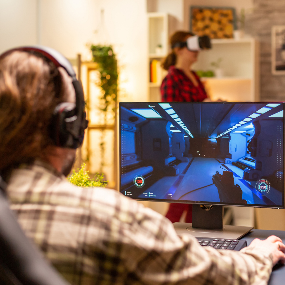 A person with headphones plays a first-person shooter game on a computer, possibly under the watch of game content moderators, while another person in the background uses a virtual reality headset. A person with headphones plays a first-person shooter game on a computer, possibly under the watch of game content moderators, while another person in the background uses a virtual reality headset.