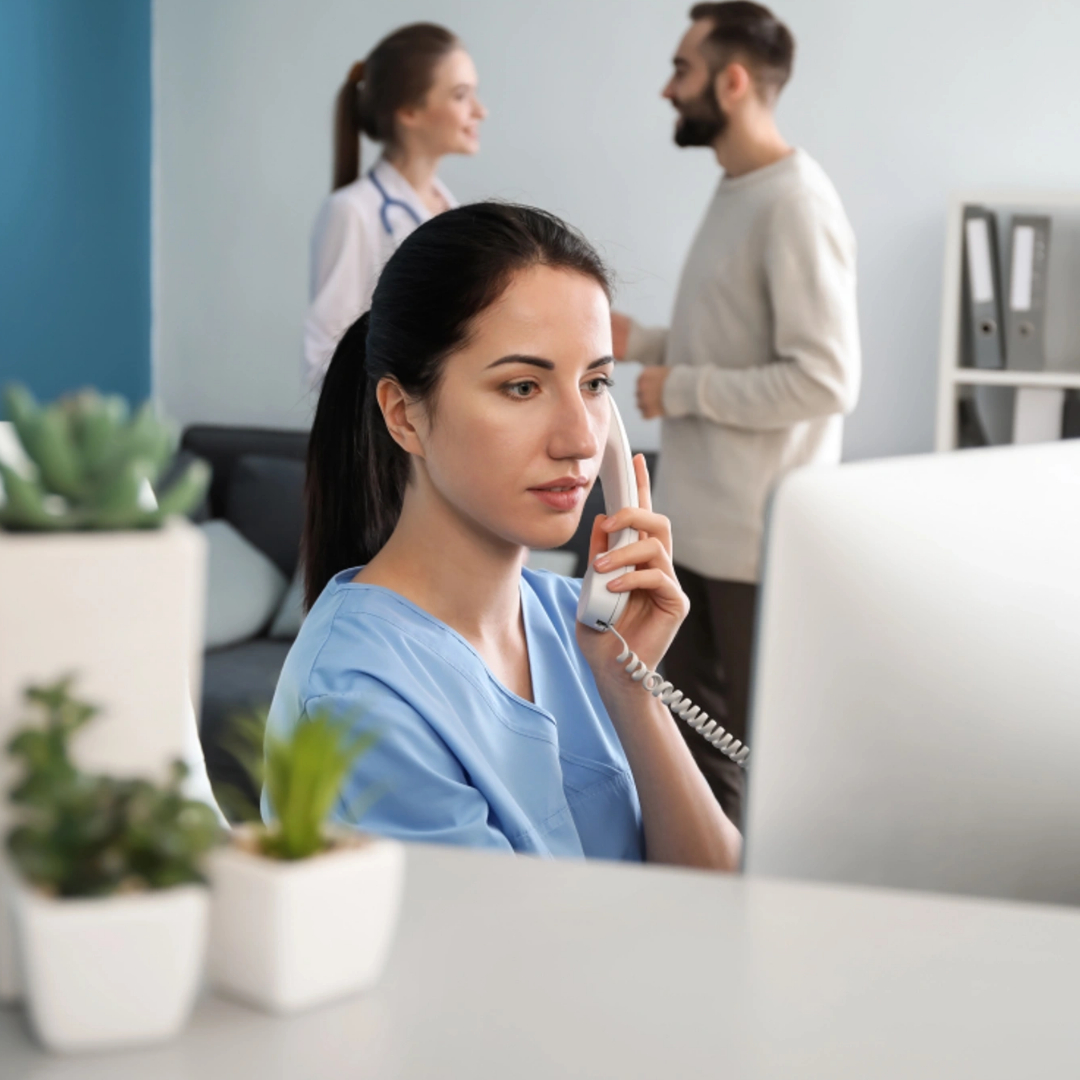A woman in medical scrubs talks on the phone at a desk with a computer, efficiently handling healthcare appointment setting, while two people converse in the background of a modern office space. A woman in medical scrubs talks on the phone at a desk with a computer, efficiently handling healthcare appointment setting, while two people converse in the background of a modern office space.