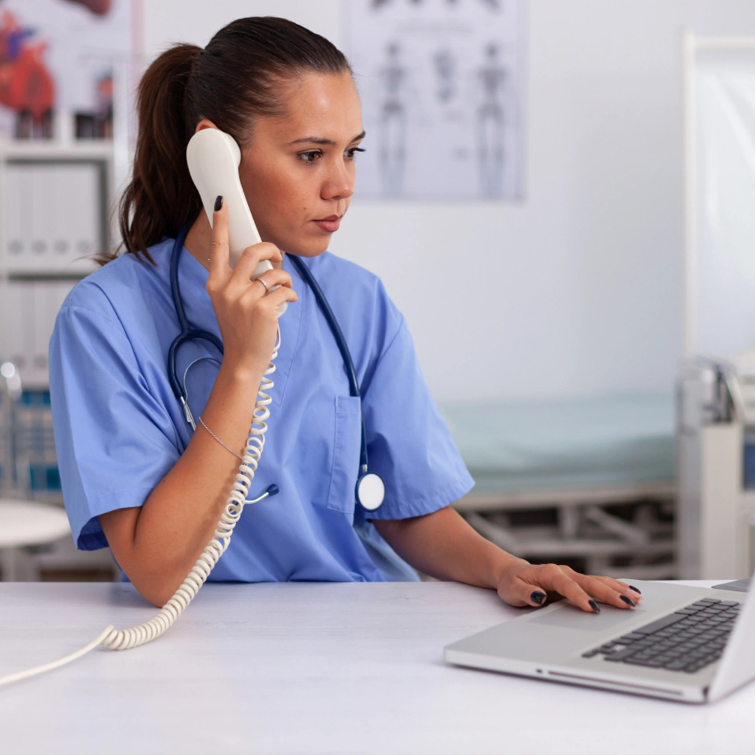Healthcare professional in scrubs sits at a desk, talking on a landline phone and using a laptop in a medical office, possibly discussing medical billing outsourcing services. Healthcare professional in scrubs sits at a desk, talking on a landline phone and using a laptop in a medical office, possibly discussing medical billing outsourcing services.